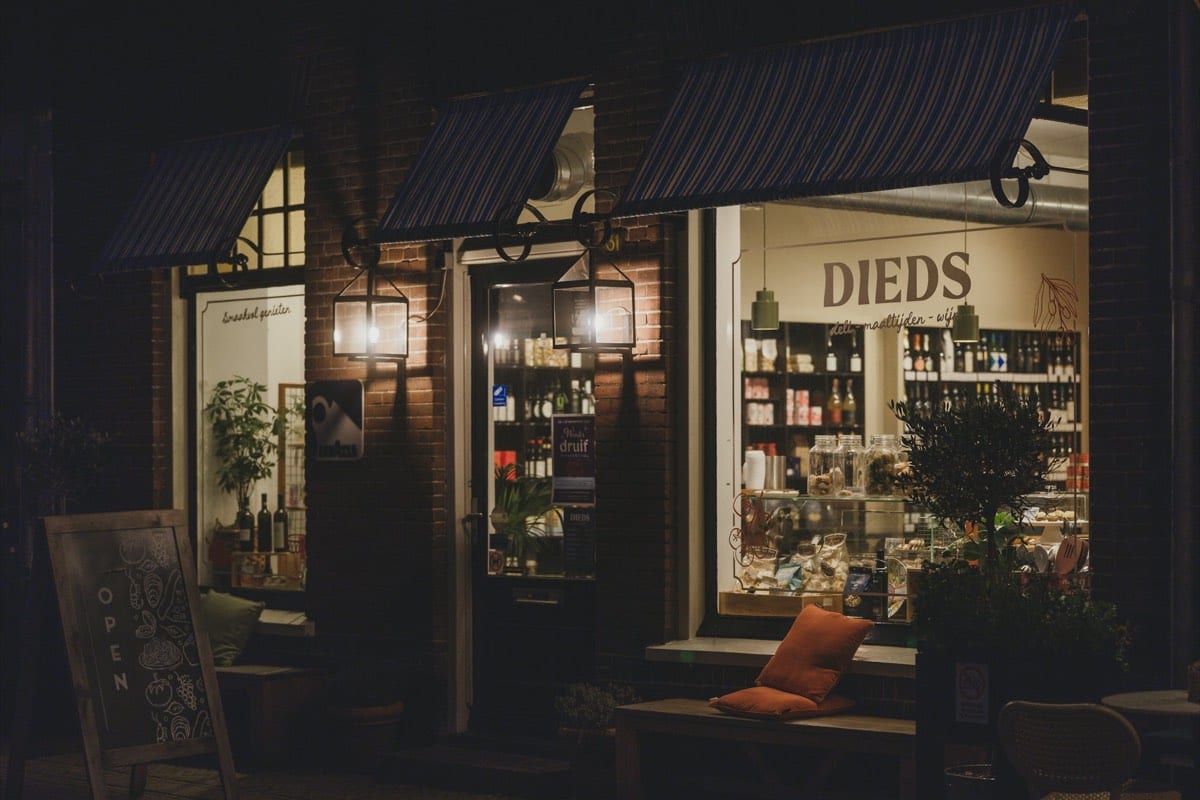 A deli and wine shop at night, its striped awnings and lit window displays glowing over a chalkboard sign on the pavement